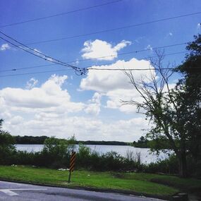 Image shows a view of a river from across a street. Above the river are blue sky, puffy clouds, and power lines.